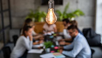 light bulb in foreground with group of people at desk blurred in background